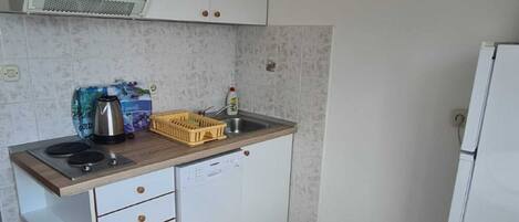 Cocina compacta con microondas, tetera y lavamanos. Hay un refrigerador pequeño y una cama con un almohadón rosa y un vaso transparente.
