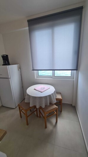 Cozy room with a round table and two chairs, featuring a small refrigerator and a window with a roller blind.