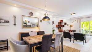 A modern living room with a wooden dining table, grey upholstered chairs, a black pendant light, and a large window with a view of greenery. - Albufeira - Anna Town (Albufeira)