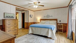 Quarto com cômoda de madeira, cama com colcha azul, ventilador de teto e mesa de cabeceira com luminária.