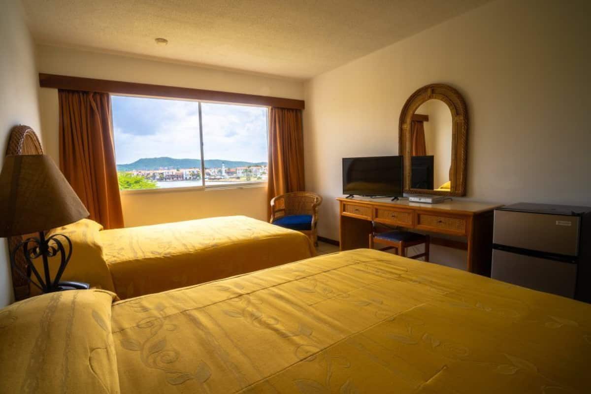 A hotel room with two beds, a desk with a television, a mirror, and a view of the city.