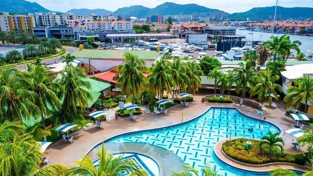 A resort with a large pool, palm trees, and a marina in the background.