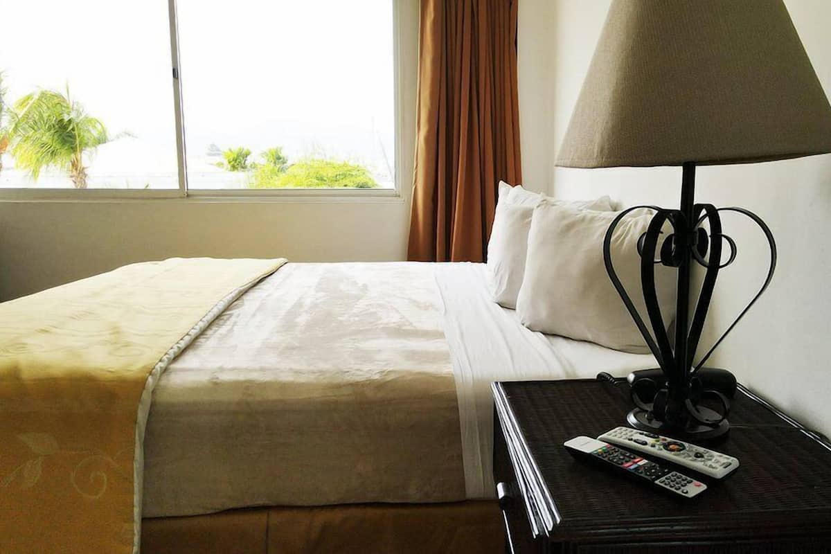 A hotel room with a bed, a bedside table with a lamp and remote controls, and a window with curtains.