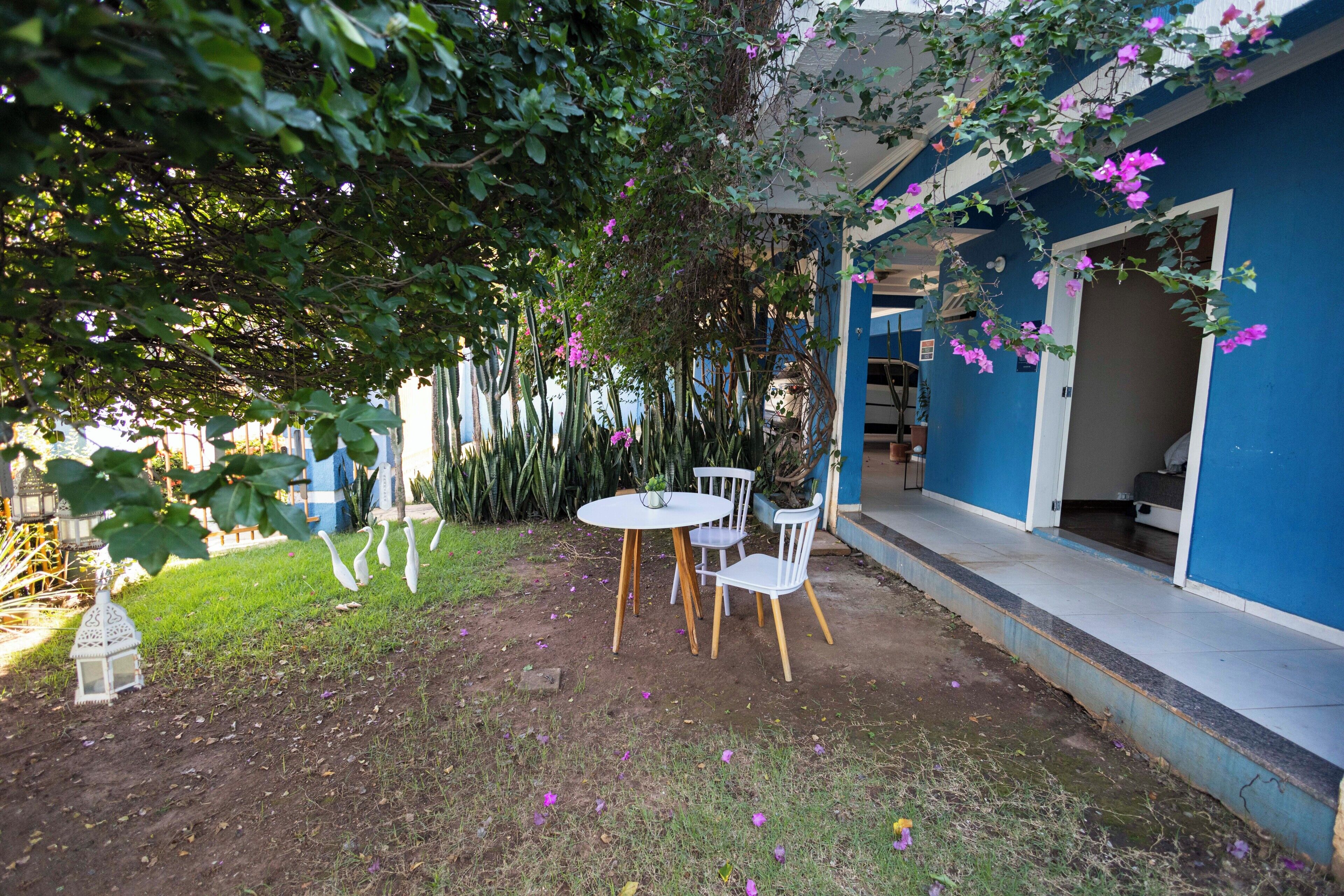 En terrasse med et bord og stole, omgivet af grøn vegetation og en blå bygning.
