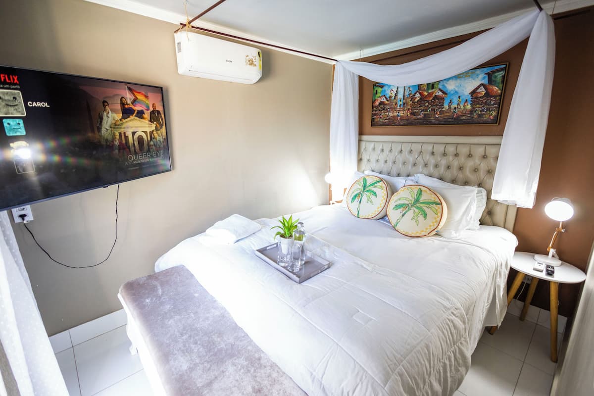 A hotel room with a large bed, a TV mounted on the wall, a painting above the bed, and a bedside table with a lamp.