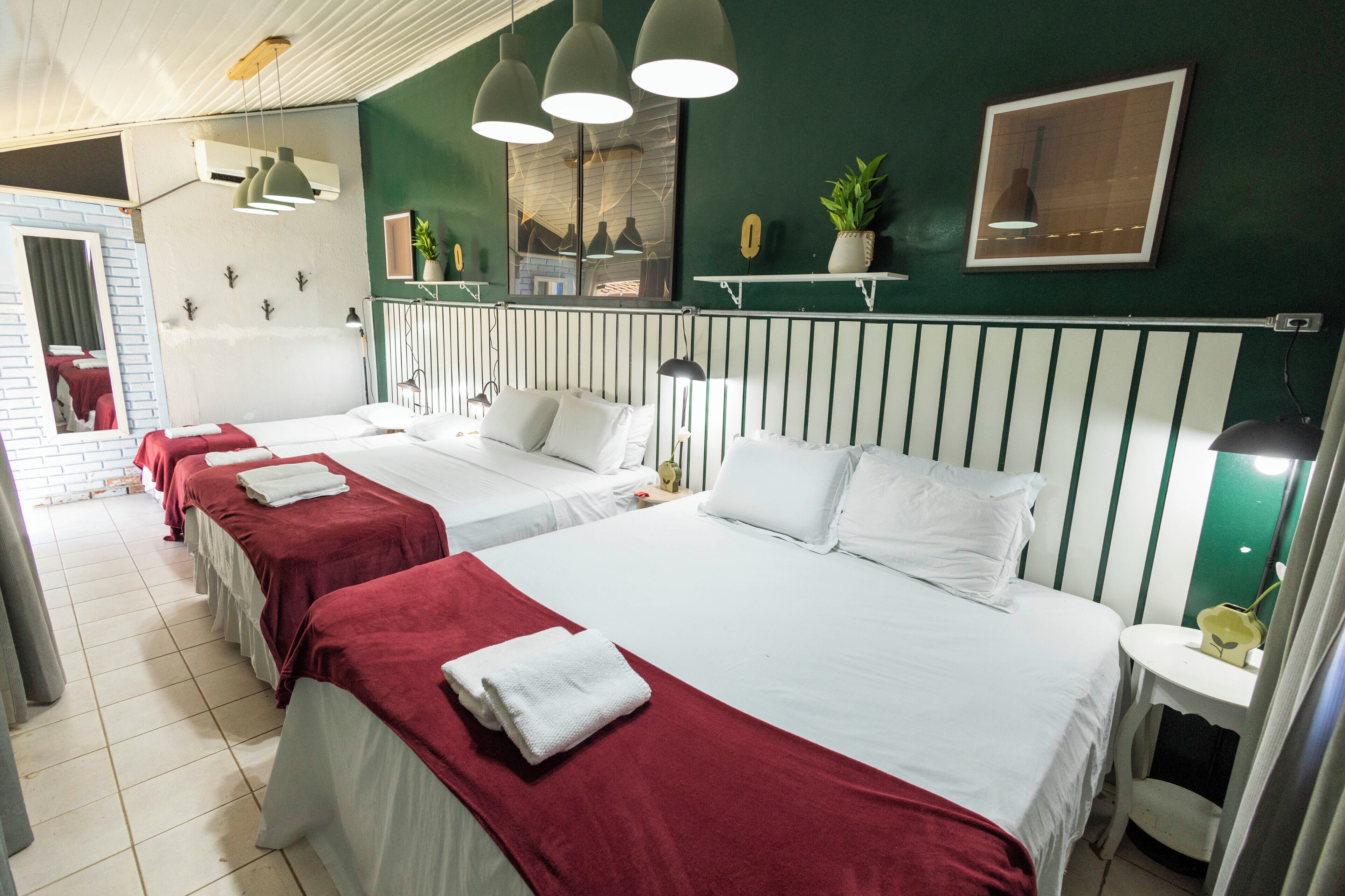 Hotellihuone, jossa on kolme sänkyä, pieni pöytä, peili ja ruukkukasvi.