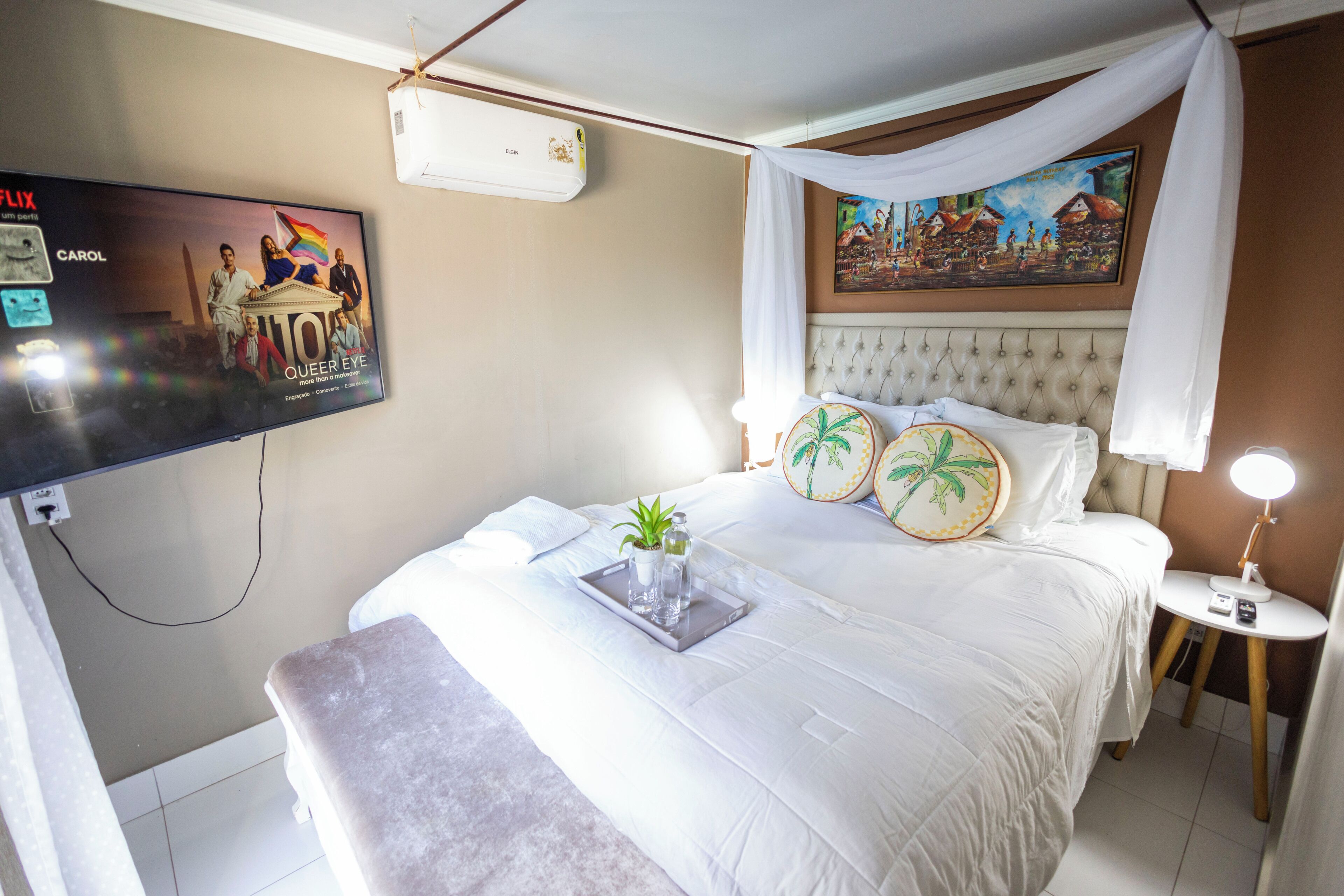A hotel room with a bed, a TV mounted on the wall, a bedside table with a lamp, and a painting above the bed.