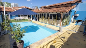 A swimming pool with a tiled deck, a patio area, and a building with a tiled roof. - Happiness House Hospedagem (Cuiaba)