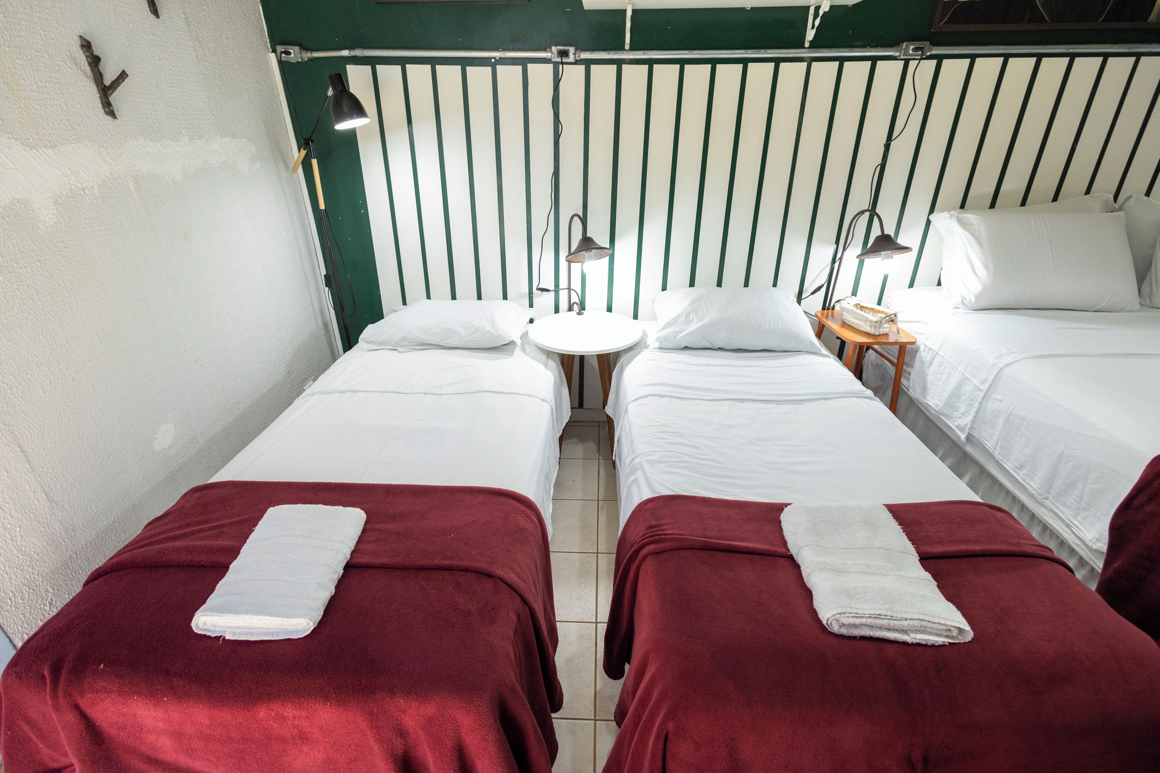 A room with two beds, each with a red blanket and a white towel, a small round table with a lamp, and a green-striped wall.