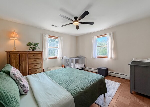 A bedroom with a bed, a crib, a ceiling fan, a dresser, and a window with curtains. - Waterfront Retreat | New Private Pier | Remodeled | Epic Sunset Views (Lexington Park)