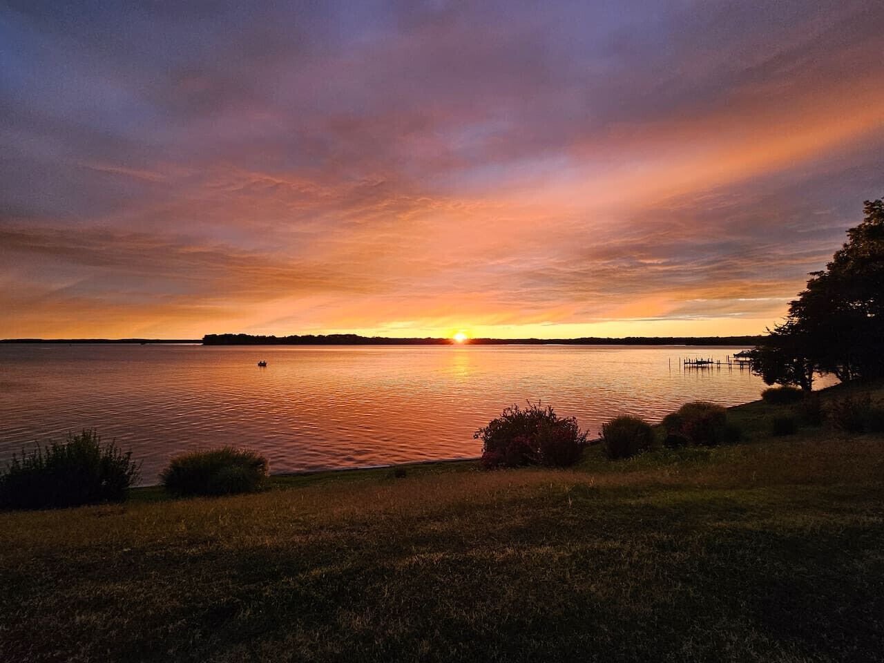 Un coucher de soleil sur un lac paisible, avec un quai et une rive bordée d’herbe.