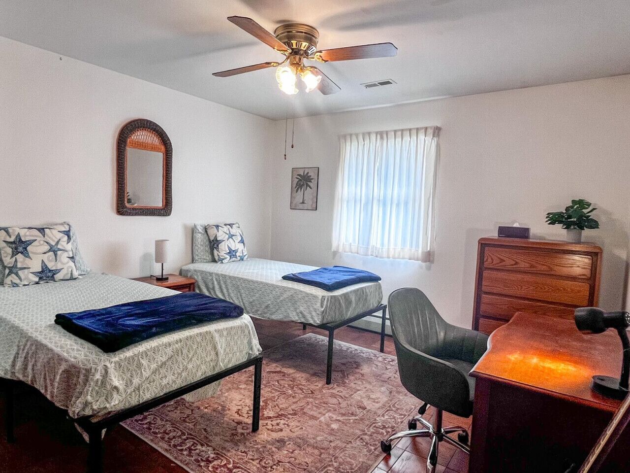 A bedroom with two beds, a desk, a chair, a ceiling fan, a dresser, and a window with curtains.