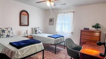 A bedroom with two beds, a desk, a chair, a ceiling fan, a dresser, and a window with curtains.
