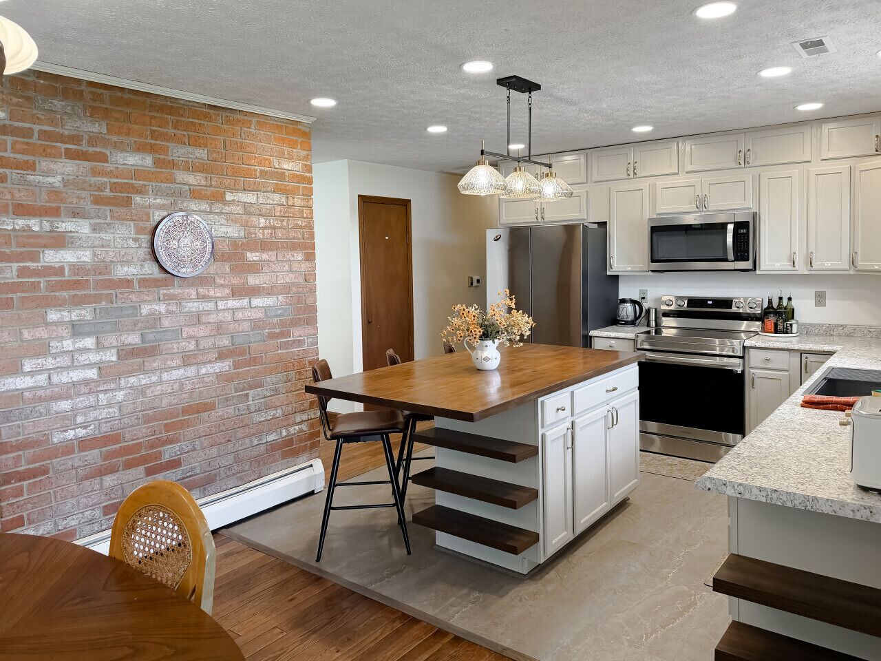 Une cuisine avec un mur en briques, une table à manger en bois, des armoires blanches, des appareils électroménagers en acier inoxydable et des luminaires suspendus.