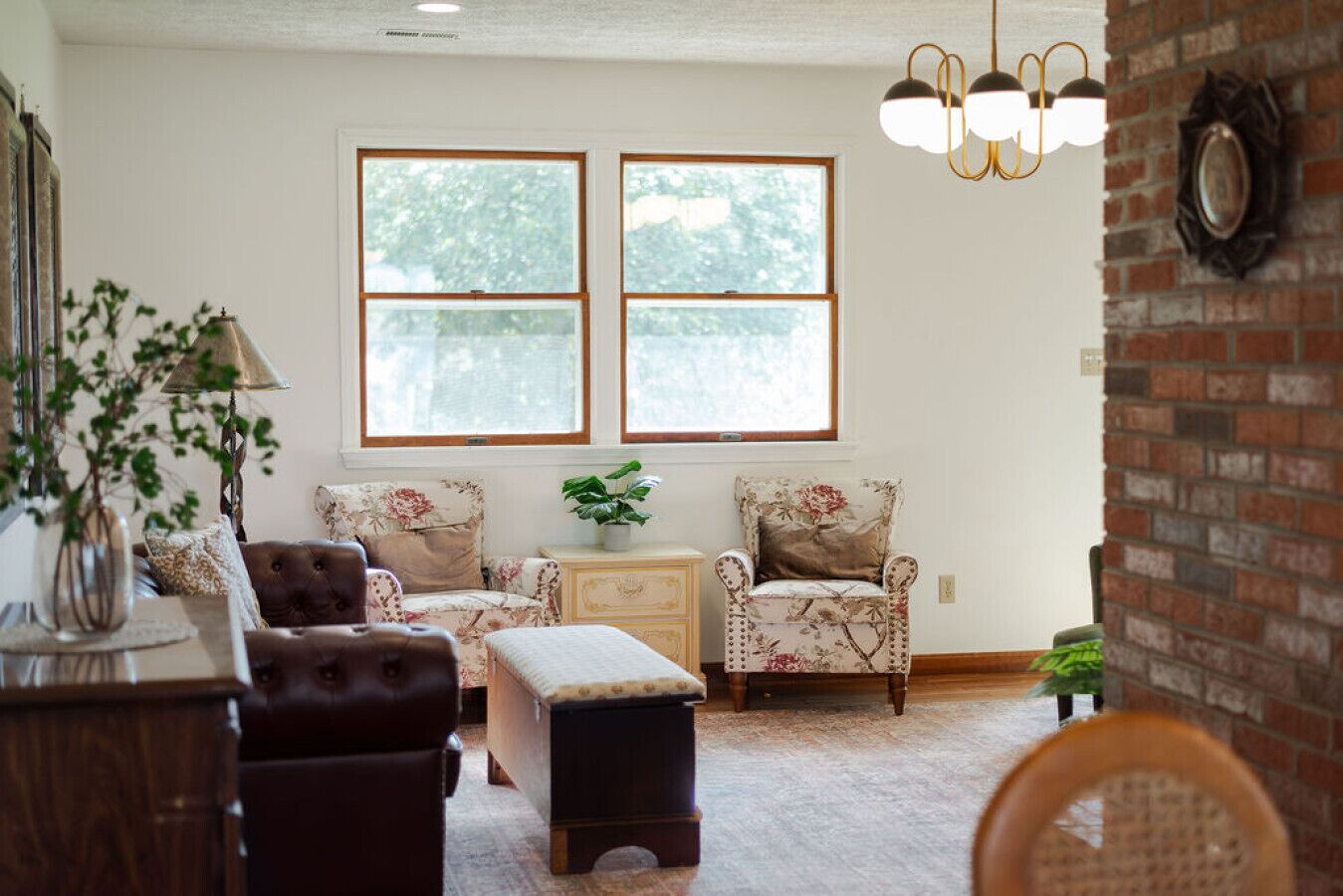 A living room with a sofa, armchair, ottoman, and side table, a window with a view, and a hanging light fixture.
