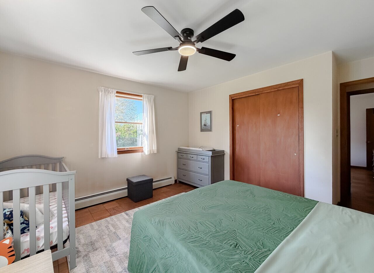 A bedroom with a bed, a crib, a dresser, a ceiling fan, and a window with curtains.