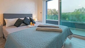 A modern bedroom with a large bed, a nightstand, and a window with a view of greenery. - Villa Fažana (Fažana)