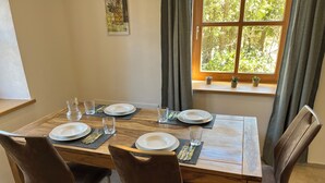 Una mesa de comedor dispuesta con platos, vasos y cubiertos, rodeada de sillas, con una ventana y cortinas al fondo.