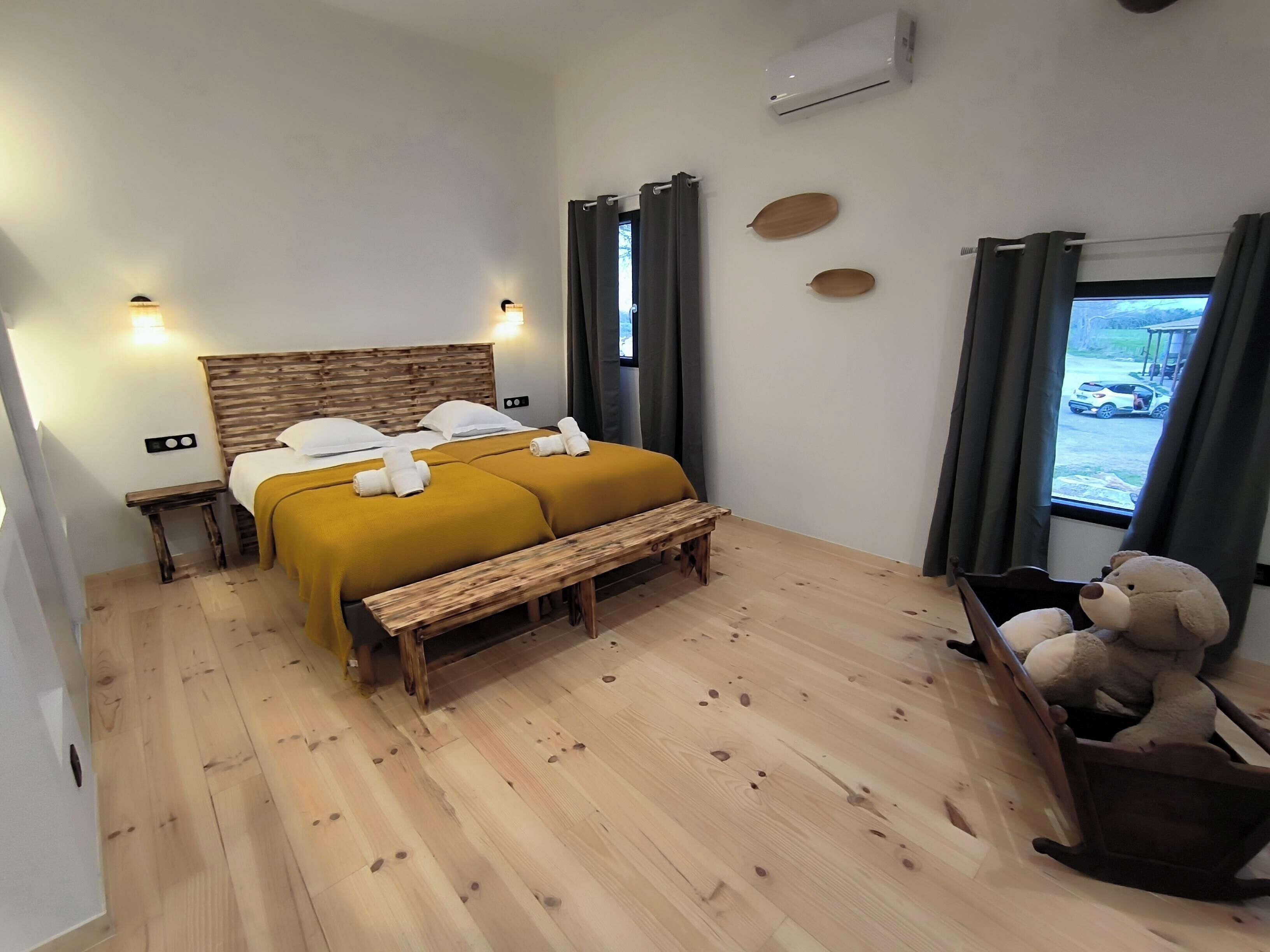 Habitación de hotel con cama de madera, dos mesitas de noche, un banco, una silla y vistas al exterior.