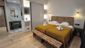 A hotel room with a wooden bench, two beds with towels, a wooden headboard, a marble wall, a mirror, and a shelf with decorative items. - Villa Blanche Domaine de la Tapie de Bouchet - in the Camargue (Arles)