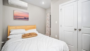 A bedroom with a wooden bed, white bedding, a painting of a sunset, a white door, and an air conditioning unit.