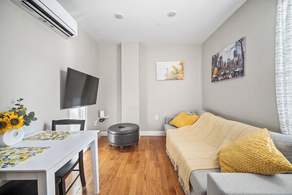 A modern living room with a flat-screen TV, a dining table with a vase of sunflowers, a grey sofa with a yellow throw, and a cityscape painting on the wall. - Lux 1st floor 3brm/1bath North End (Boston)