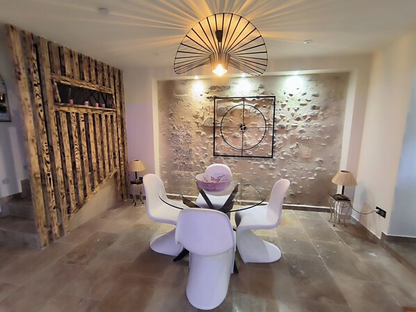 A dining area with a round table and four white chairs, a wooden wall with a rustic design, a modern light fixture, and a textured wall with a circular design. - Villa Framboise Domaine de la tapie de Bouchet - en Carmargue (Arles)
