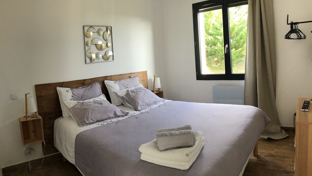 A neatly made bed with a grey bedspread and pillows, a wooden headboard, a bedside table with a lamp, and a window with curtains. - Gite for 2, in the countryside, 15 minutes from the medieval town of Carcassonne (Verzeille)