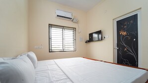 A bedroom with a bed, an air conditioner, a window with bars, a door with a floral design, and a shelf with a television.