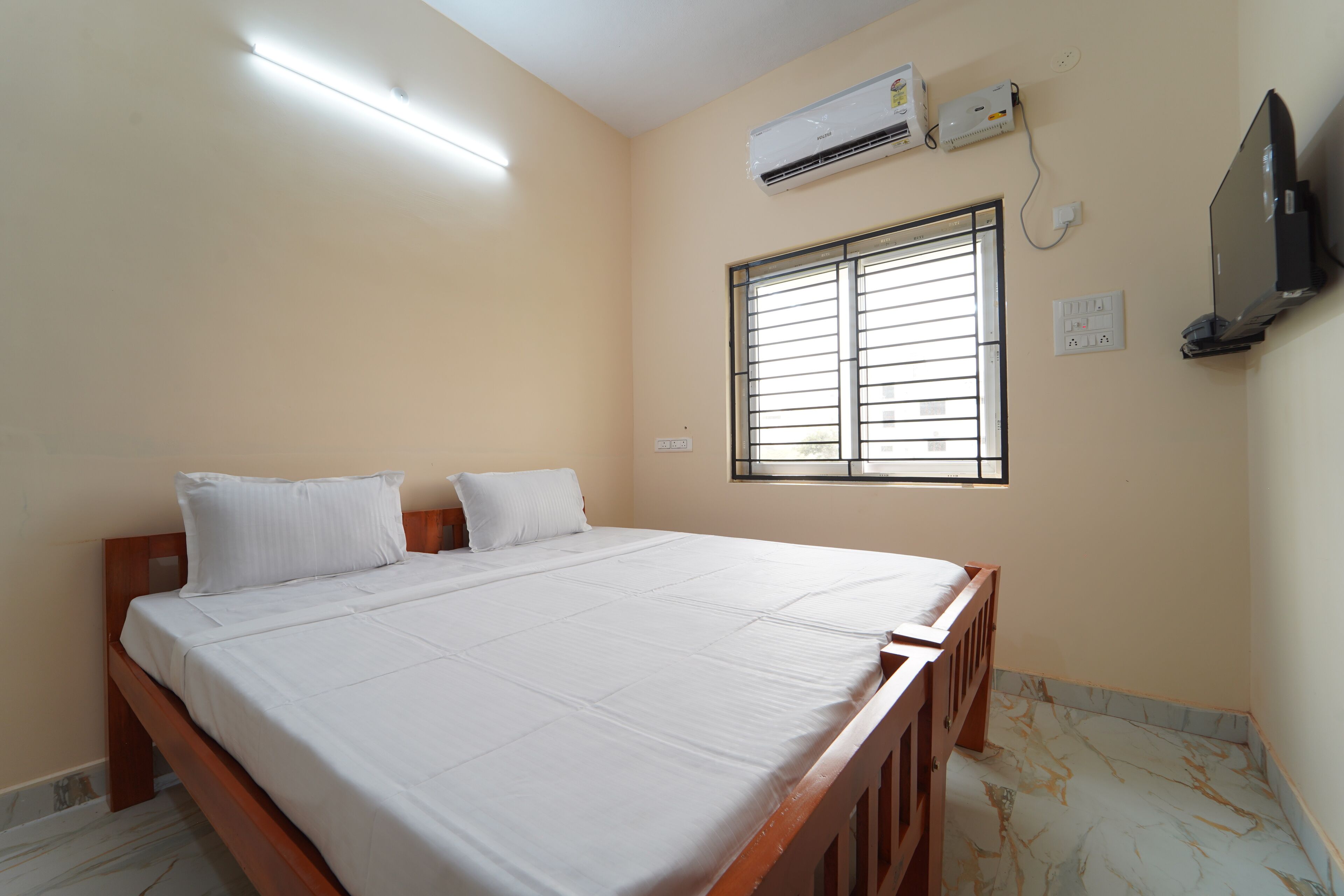 A bedroom with a large bed, a window with blinds, an air conditioning unit, and a television mounted on the wall.