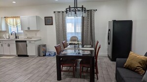 A modern kitchen with white cabinets, a dishwasher, and a refrigerator. There is a dining table with chairs and a chandelier above it.