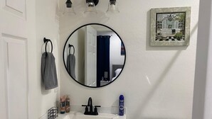 A bathroom with a white sink, a round mirror, and a toilet. There are towels hanging on hooks, a framed picture on the wall, and a small shelf with toiletries.