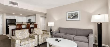 A living room with a grey sofa, patterned armchairs, a white coffee table, and a floor lamp. There is a kitchen area in the background with white cabinets and a black refrigerator.