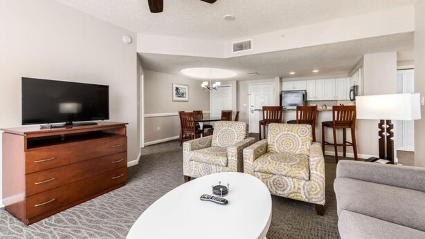 Habitación de hotel con televisión, cómoda de madera, dos sillones estampados, una mesa de centro blanca y un área de comedor con sillas y una mesa.