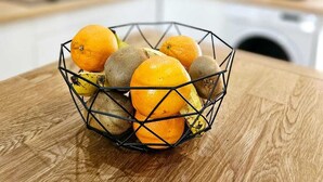 Eine Küche mit einem Holztisch, auf dem sich ein Drahtkorb voller Orangen und Kiwis befindet.