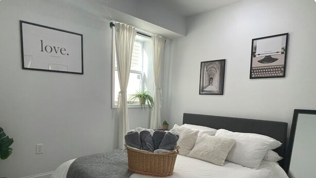 A neatly arranged bedroom with a bed, a basket of towels, a framed word on the wall, and a window with curtains.