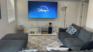 A living room with a flat-screen TV displaying the Disney+ logo, a grey sofa, a coffee table with decorative items, and a patterned rug.
