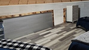 Une pièce avec un plafond en pente, un ventilateur de plafond, un lit recouvert d’une couverture à carreaux et une commode.