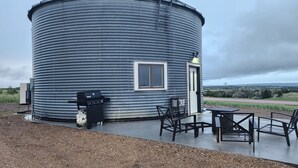 Un bâtiment cylindrique en métal, doté d’une porte et d’une fenêtre, avec un espace extérieur aménagé avec des chaises et des tables, ainsi qu’un barbecue.