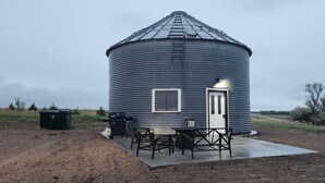 Un bâtiment cylindrique en métal, doté d’une porte et de fenêtres, entouré d’une terrasse en béton aménagée avec du mobilier d’extérieur.