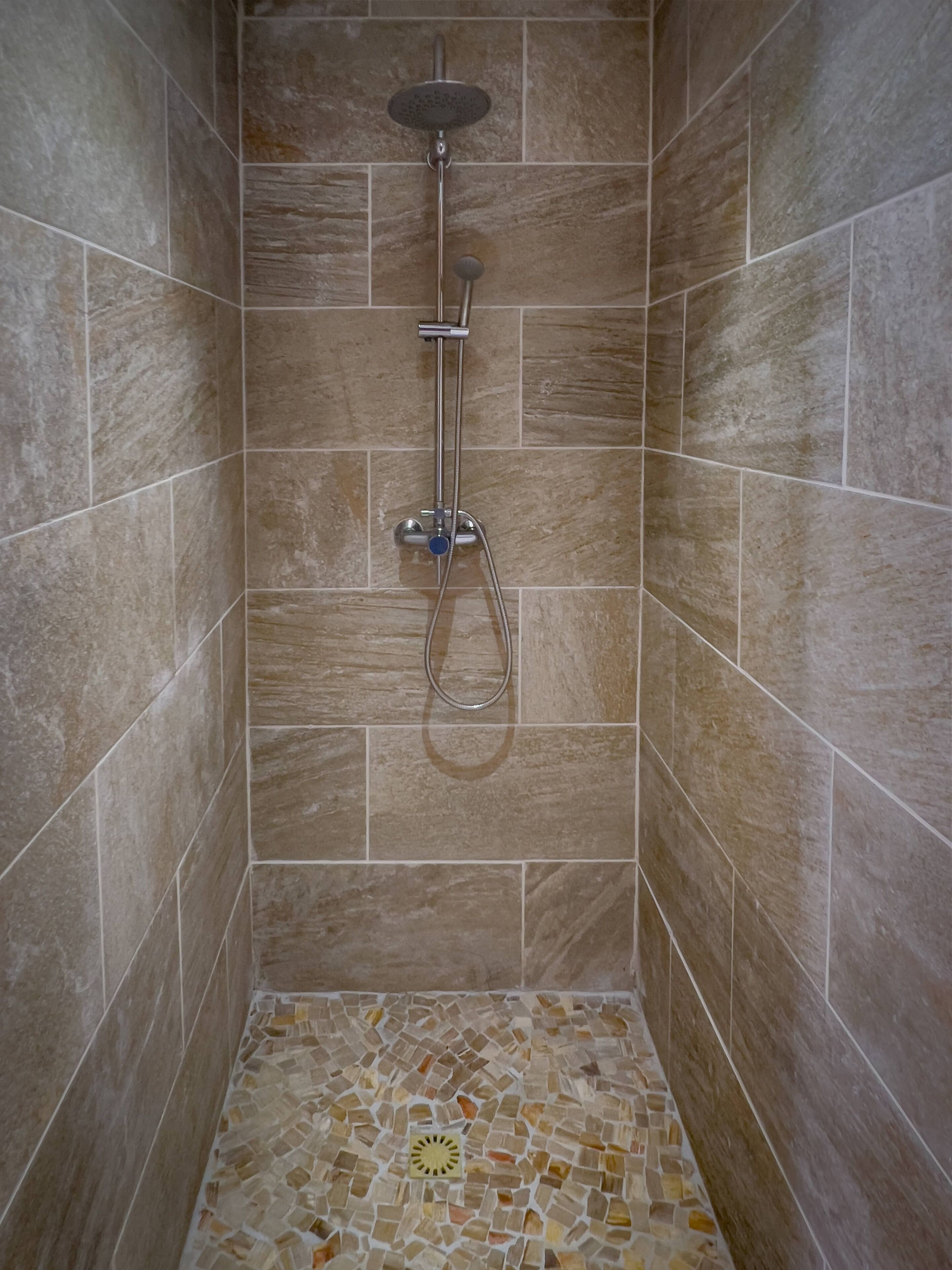 A shower with a rain showerhead and handheld showerhead, tiled walls, and a mosaic floor.