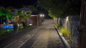 Vue nocturne d’une allée menant à une maison, avec une piscine, une maison en bois et un mur de pierres illuminé.
