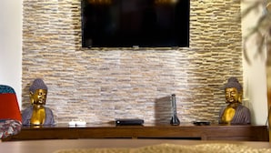 Un salon avec un mur en pierre, un téléviseur à écran plat fixé au-dessus d’une console en bois, et deux statues de Bouddha.