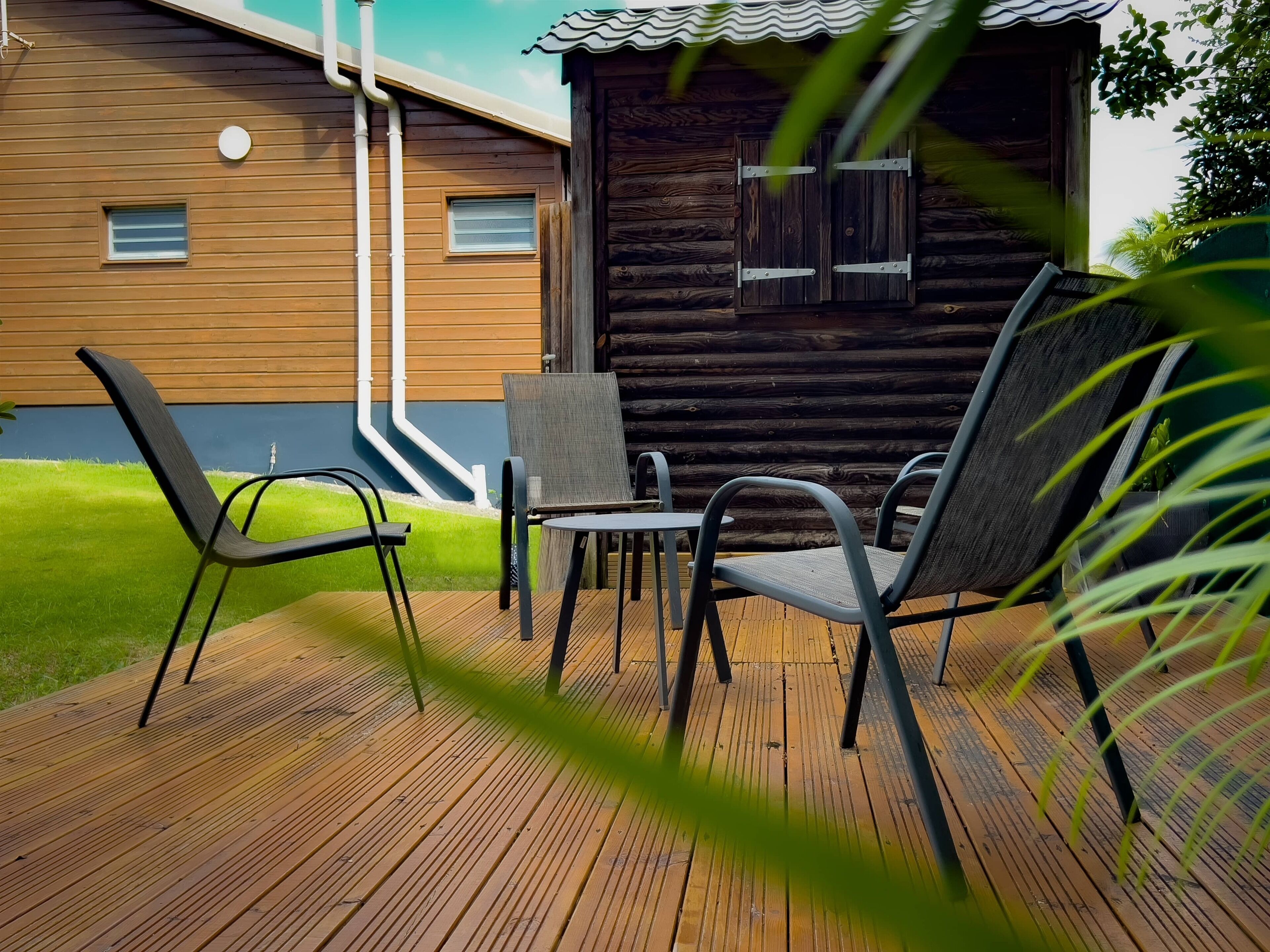 Une terrasse en bois avec deux chaises et une table, une petite fenêtre et, au fond, une cabane en bois.
