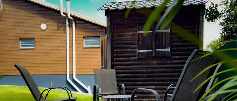 Une terrasse en bois avec deux chaises et une table, une petite fenêtre et, au fond, une cabane en bois.