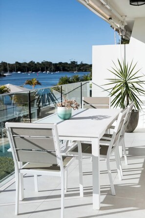 A white outdoor dining area with a view of the sea, a potted plant, and a glass railing.