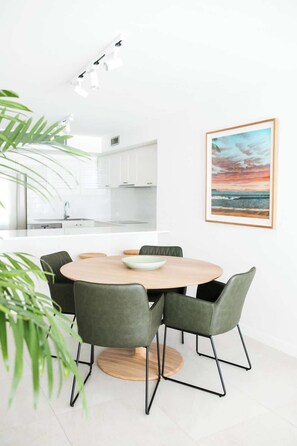 Uma cozinha moderna com armários brancos, uma área de jantar com mesa redonda e quatro cadeiras, e um quadro com uma pintura de pôr do sol na parede.