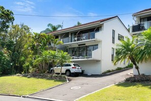 A two-story residential building with a balcony, a car parked in front, and surrounding greenery.