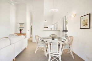 Um interior moderno com área de jantar, cadeiras de vime e mesa de vidro, paredes brancas e ventilador de teto.