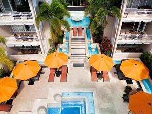 A hotel pool area with multiple lounge chairs and umbrellas, surrounded by palm trees and a central water feature.